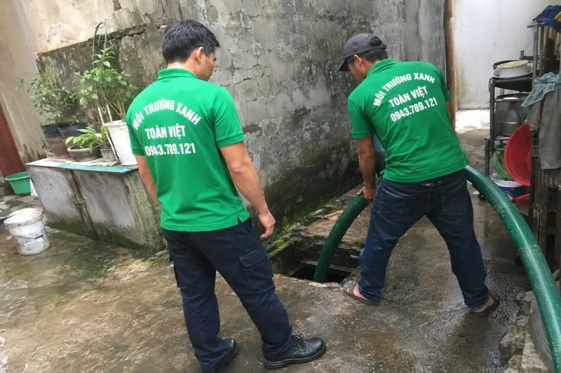 Hai nh&acirc;n vi&ecirc;n &aacute;o xanh bố tr&iacute; đường ống h&uacute;t xuống một hố mở ngo&agrave;i s&acirc;n hẹp.